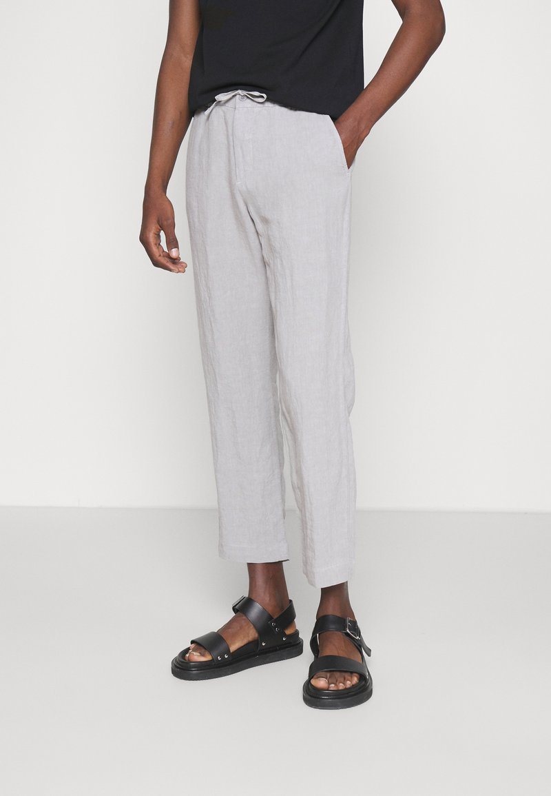 Osoba ubrana w jasnoszare lniane spodnie z wiązaniem w pasie oraz czarne skórzane sandały, stojąca na tle jednego białego tła.