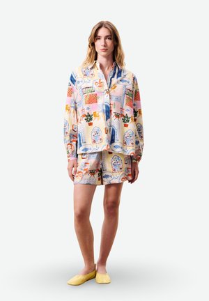 Femme debout portant une chemise colorée à motifs boutonnée et un short assorti avec des illustrations de chaises, plantes et fenêtres, associée à des chaussures jaunes.
