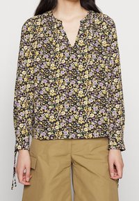 Blomsterlangærmet bluse med sort baggrund, der har gule, lilla og hvide blomster. V-hals og samlede ærmer med bindebånd.