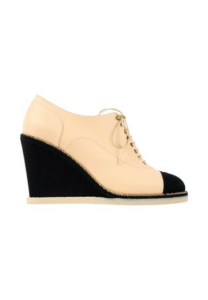 Chaussure compensée en cuir beige avec embout et talon noirs, fermeture à lacets et garniture décorative en chaîne dorée.