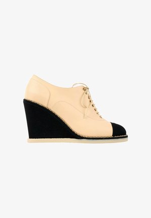 Chaussure compensée en cuir beige avec embout et talon noirs, fermeture à lacets et garniture décorative en chaîne dorée.
