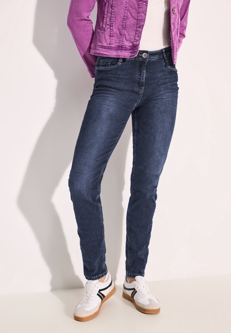 Jeans ajustados de color azul oscuro con un diseño de cinco bolsillos y un desvanecimiento sutil. Combinados con zapatillas blancas con acentos negros.