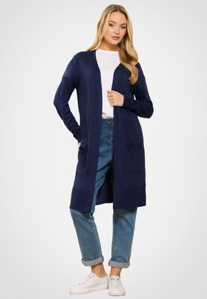 Kvinde iført en navyblå lang cardigan, hvid skjorte, op rullede blå jeans og hvide sneakers, stående med den ene hånd i lommen.