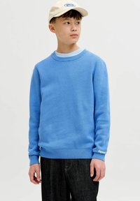 Garçon portant un pull bleu clair, un jean noir et une casquette blanche, regardant sur le côté sur un fond blanc uni.