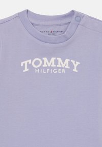 Lys lilla bomulls T-skjorte med rund hals, med hvit "TOMMY HILFIGER" logo trykt over brystet og trykknapp på skulderen.