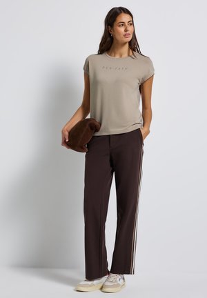 Beige short-sleeved t-shirt with "NEW PATH" text, paired with dark brown trousers featuring white side stripes, and light-colored sneakers.