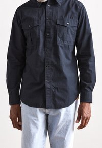 Camisa abotonada de marina hecha de algodón, con dos bolsillos en el pecho y un dobladillo redondeado, combinada con unos jeans de color azul claro.