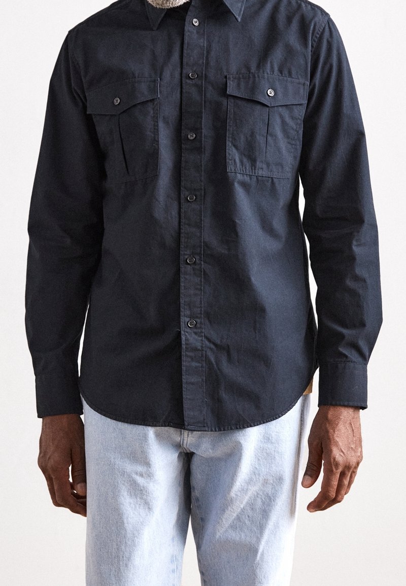 Camisa abotonada de marina hecha de algodón, con dos bolsillos en el pecho y un dobladillo redondeado, combinada con unos jeans de color azul claro.