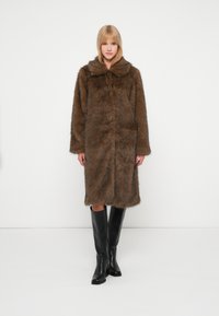 Brūns mākslīgā kažokādas mētelis ar plašu apkakli, garām piedurknēm un vidēja garuma dizainu. Valkāts virs ceļgala melnām zābakiem. Gluda tekstūra.