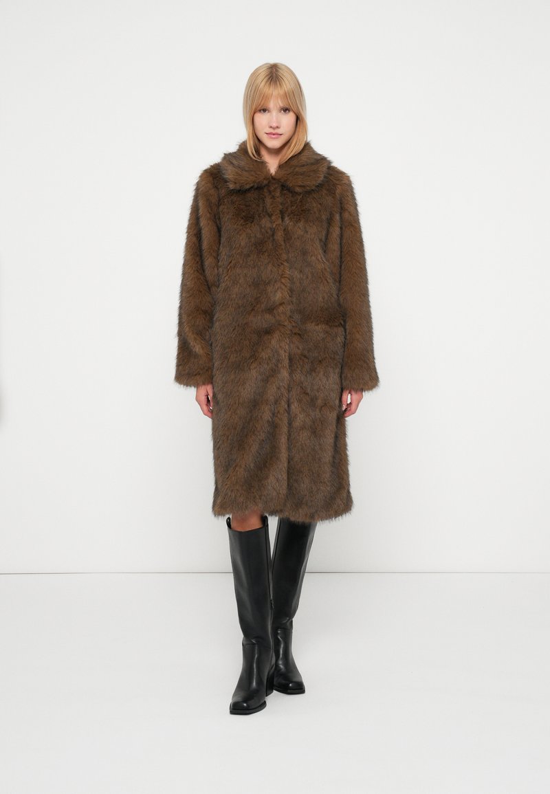 Brūns mākslīgā kažokādas mētelis ar plašu apkakli, garām piedurknēm un vidēja garuma dizainu. Valkāts virs ceļgala melnām zābakiem. Gluda tekstūra.