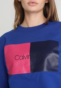 Modrá mikina s kulatým výstřihem, která vyniká obdélníkovým barevným blokem v růžové a námořnické modři, uprostřed je lesklé černé logo "Calvin Klein".
