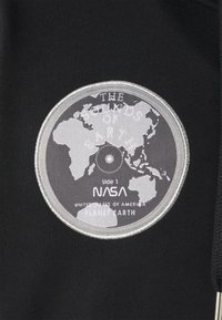 Tela negra con un parche bordado redondo en plata que muestra un mapa del mundo y el texto: "LOS SONIDOS DE LA TIERRA," "NASA," y "PLANETA TIERRA."