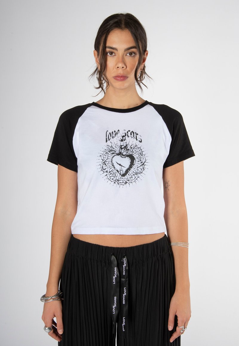 Camiseta recortada con un cuerpo blanco y mangas raglán negras, con un diseño central de corazón negro impreso y texto decorativo arriba.
