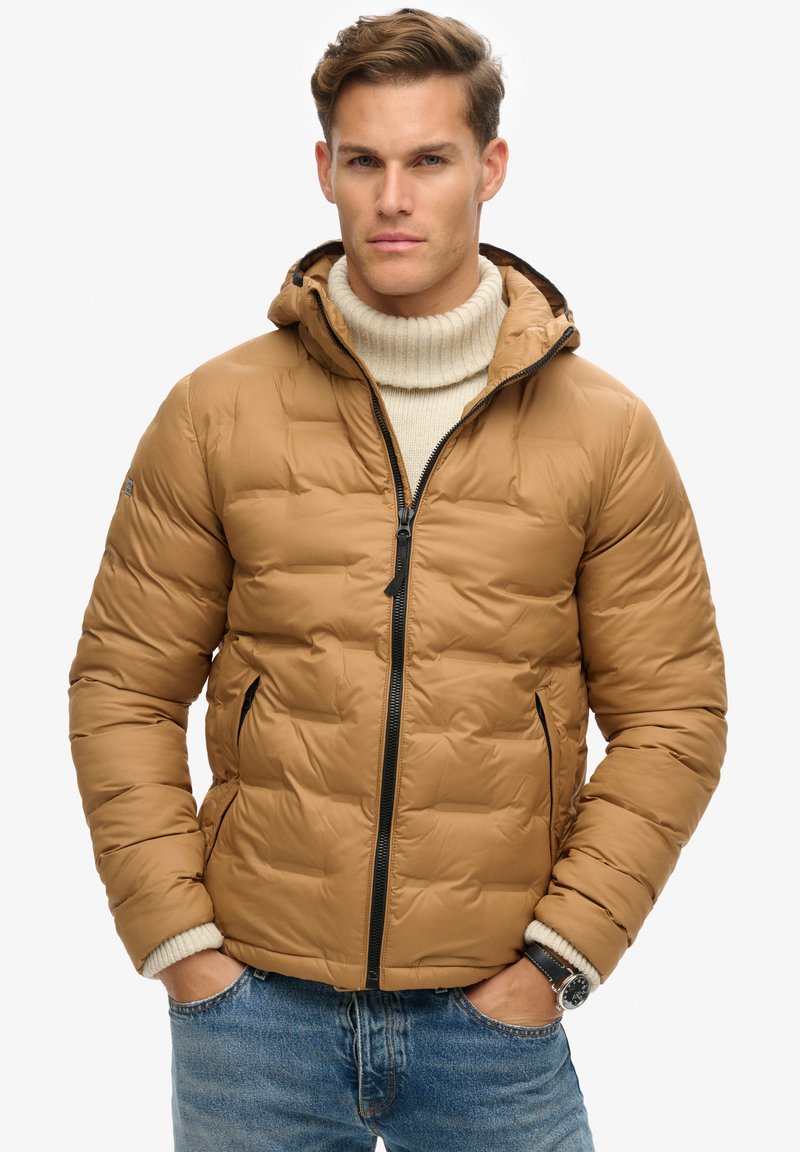 Bruine pufferjack met een capuchon, ritssluiting en elastische manchetten. Kenmerkt zich door een gestikte uitvoering en zijzakken, gedragen over een crèmekleurige coltrui.