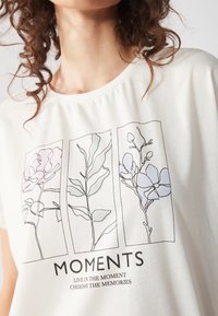 Heled värvi puuvillane T-särk, millel on botaanilised joonistused pastelltoonides, koos sõnaga "MOMENTS" ja sloganiga all.