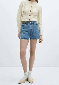 Cremfarbener Grobstrick-Cardigan mit braunen Knöpfen, kombiniert mit Jeansshorts. Das Modell trägt creefarbene Socken und beige Ballerinas. Neutraler Hintergrund.