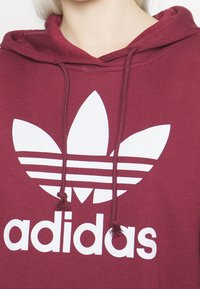 Felpa con cappuccio color bordeaux con logo bianco adidas che presenta tre foglie e tre strisce orizzontali. Realizzata in tessuto testurizzato con lacci.