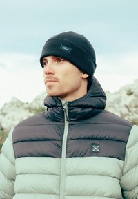 Bonnet en tricot noir et une veste matelassée bicolore avec une partie supérieure gris foncé et une partie inférieure vert clair. Comprend une fermeture éclair à l'avant et une patte avec logo.