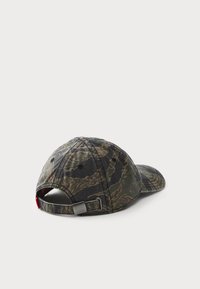 Kamuflasje caps laget av stoff, med buet skygge, justerbar stropp bak, og fremtredende sømmer i svart og grønt.