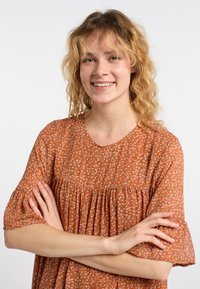 Roestkleurige blouse met korte, gerimpelde mouwen; heeft een bloemenpatroon met kleine witte, roze en blauwe details; losse pasvorm.