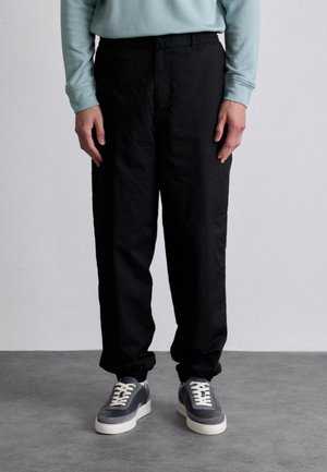 Black, textured trousers with a loose fit, featuring an elastic waistband. Paired with gray sneakers, displaying a rounded toe and white laces.