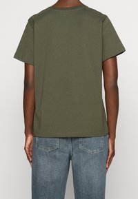 Personne portant un t-shirt à manches courtes vert olive et un jean en denim bleu, vue de dos sur un fond uni.