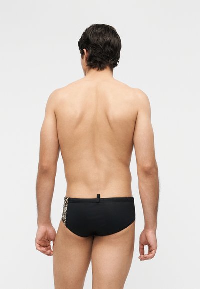 Άνδρας με σκουρόχρωμα μαλλιά στέκεται με την πλάτη γυρισμένη, φορώντας μαύρα μαγιό σλιπ με λεπτομέρεια λεοπάρ στην μία πλευρά, μπροστά σε ένα απλό φόντο.