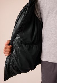 Veste isolée noire avec une finition brillante, dotée d'une poche intérieure, d'une fermeture éclair et d'un design léger.
