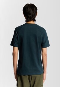 Mørkegrøn t-shirt i bomuld med rund hals og korte raglanærmer, der har et lille logo på ryggen nær halsen.