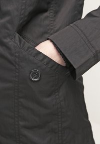 Svart, vattenavvisande jacka med en mjuk textur, med en knappdetalj och sidfickor. Närbild på ärm och handled.