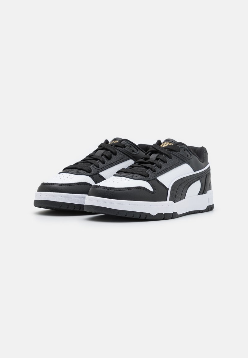 Chaussures de sport noires et blanches avec un dessus en cuir lisse, un bout rond, un laçage proéminent et une semelle en caoutchouc texturée. Détail de marquage doré visible.