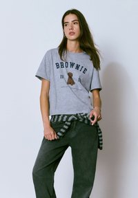 Camiseta de algodón gris con el texto "BROWNIE", un gráfico de un oso y un corte relajado. Combinada con pantalones oscuros y un suéter a rayas atado alrededor de la cintura.
