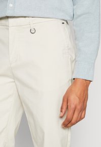 Pantaloni beige chiaro realizzati in misto cotone, con una texture liscia, dettaglio ad anello metallico in vita e tasche laterali standard.