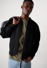 Black bomber jacket with a grey zipper, ribbed cuffs, and a relaxed fit, worn over an olive green shirt, with light blue jeans.