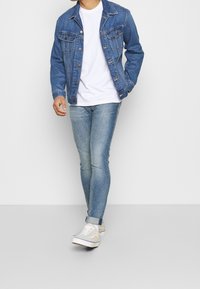 Chaqueta de mezclilla con dos bolsillos en el pecho, jeans ajustados de color azul claro y zapatillas grises con cordones blancos, llevados sobre una camiseta blanca.