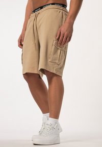 Shorts cargo beiges avec une taille élastique, un cordon de serrage et deux poches latérales. Portés avec des baskets montantes blanches et des chaussettes mi-mollet.