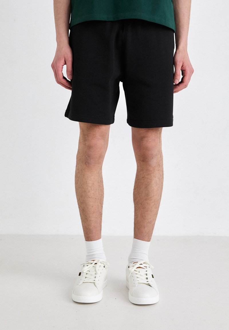 Pantalones cortos de algodón negros con cintura elástica, corte recto y zapatillas blancas de contraste. Calcetines blancos visibles por encima del tobillo.