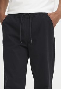 Svarte bukser med elastisk midjebånd og snor. Har sidelommer og løs passform. Glatt stoff uten synlige mønstre.