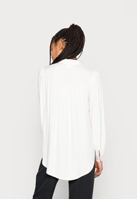 Vit långärmad blus med veckad baksida, böjd fåll och knäppte cuffs. Tillverkad av ett lätt tyg med en slät yta.