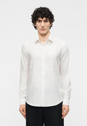 Mann mit lockigem Haar trägt ein weißes, geknöpftes Langarmhemd und schwarze Hose, steht vor einem schlichten hellen Hintergrund.