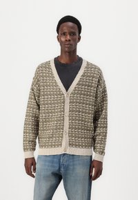 Strikket cardigan i grøn og creme med et tekstureret mønster. Har V-hals, lange ærmer og knapper foran, parret med lyse denim jeans.