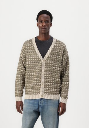 Gilet en tricot vert et crème avec un motif texturé. Présente un col en V, des manches longues, et des boutons sur le devant, associé à un jean en denim clair.