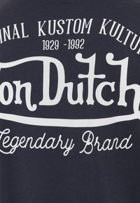 Logo de Von Dutch con el texto "Original Kustom Kulture 1929-1992" y "Legendary Brand" en blanco sobre un fondo de tela oscura.