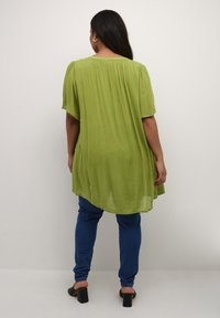 Tunic verde a maniche corte con vestibilità ampia e orlo arrotondato. Indossata sopra jeans blu, realizzata in un tessuto leggero e testurizzato.