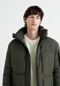 Mørkegrøn pufferjakke med høj krave og trykknaplukning, parret med en cremefarvet strikket sweater. Blød tekstur, quiltet design.