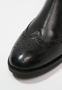 Zapato de cuero negro con punta redondeada, detalles de brogue y una textura suave y brillante. Presenta perforaciones intrincadas y un tacón bajo.