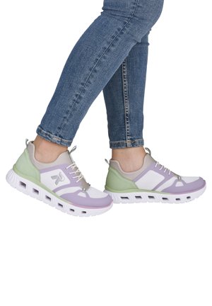 Person trägt weiße, lavendelfarbene und grüne Sportschuhe mit belüfteten Sohlen und blaue Skinny-Jeans und posiert vor weißem Hintergrund.