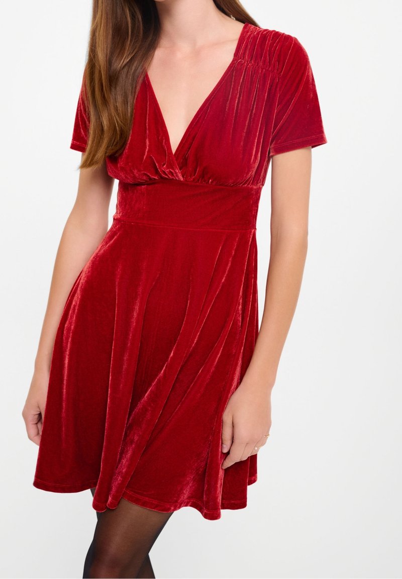 Femme portant une robe courte en velours rouge avec un col en V profond et des épaules froncées, debout devant un fond blanc.