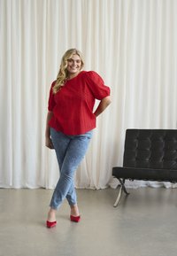Femme souriante se tient les jambes croisées, portant un blouse rouge, un jean bleu et des talons rouges, dans une pièce lumineuse avec des rideaux blancs et un banc noir.
