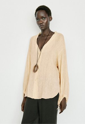 Donna che indossa una camicetta beige morbida e strutturata e pantaloni neri a gamba larga, accessoriata con una lunga collana con un grande pendente circolare.